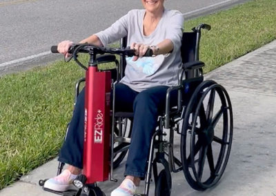 An image of an older woman sitting in a manual wheelchair with the EZRide unit connected. She is riding down a neighborhood sidewalk