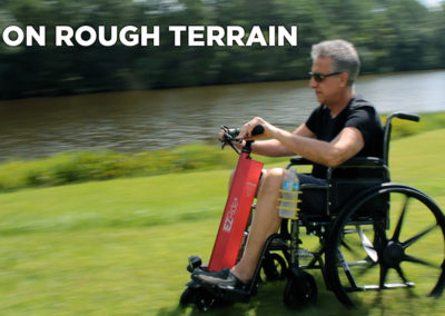 An image of a man sitting in a manual wheelchair connected to the EZRide unit, moving at speed over grassy terrain beside water. The text states "EZ on rough terrain"