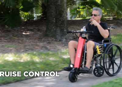 An image of an older man riding in a manual wheelchair with the EZRide connected, traveling down a sidewalk while holding a drink and smoking a cigar. The man is not using his hands on the controls, relying on the EZRide cruise control