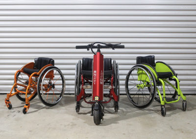 An image of three WCMX rigid frame wheelchairs - burnt orange, red, and lime green. The red wheelchair has the EZRide unit connected