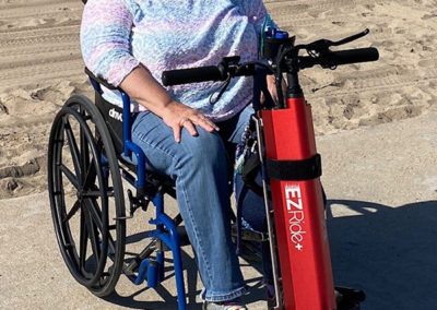 An image of an EZRide+ owner sitting in her wheelchair, connected to her EZRide+ in front of a sandy beach, on a concrete sidewalk